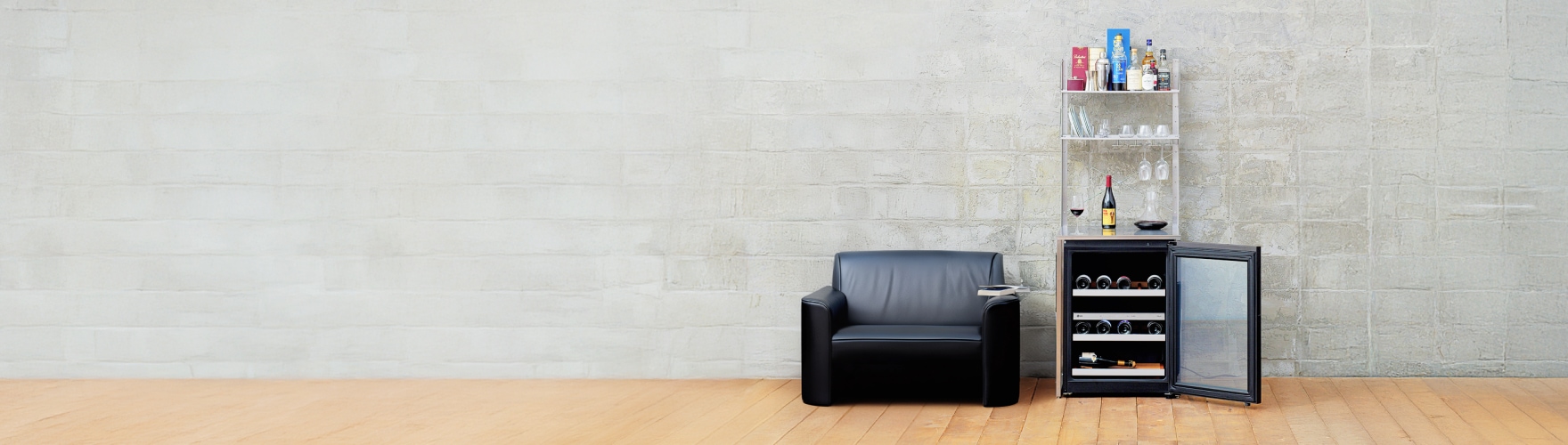 콘크리트 질감의 벽 앞에 검은색 1인용 소파와 유리문이 열린 와인셀러장이 나란히 놓여 있다. 와인셀러장 위 선반에는 와인잔과 주류 병들이 정리되어 있다.