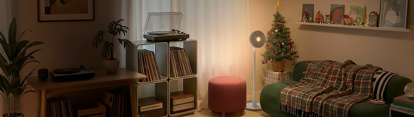 크리스마스 장식으로 아늑하게 꾸며진 거실 전경입니다. 왼쪽에는 턴테이블과 LP가 정리된 선반이 있으며, 오른쪽에는 크리스마스 트리와 스탠드 조명이 자리하고 있습니다. 초록색 소파에는 체크 무늬 담요가 펼쳐져 있고, 벽 선반에는 그림과 소품이 진열되어 있습니다.
