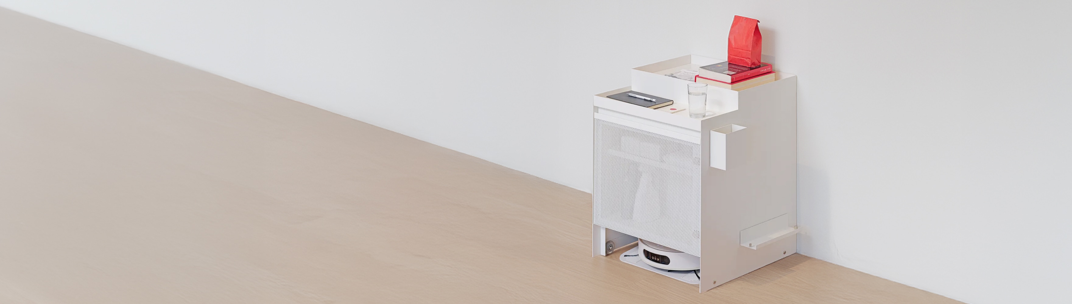 흰색 미니멀 수납 가구, 레어로우 위에 겁과 노트, 빨간 봉투가 놓여 있고 하단엔 로봇청소기가 있다.