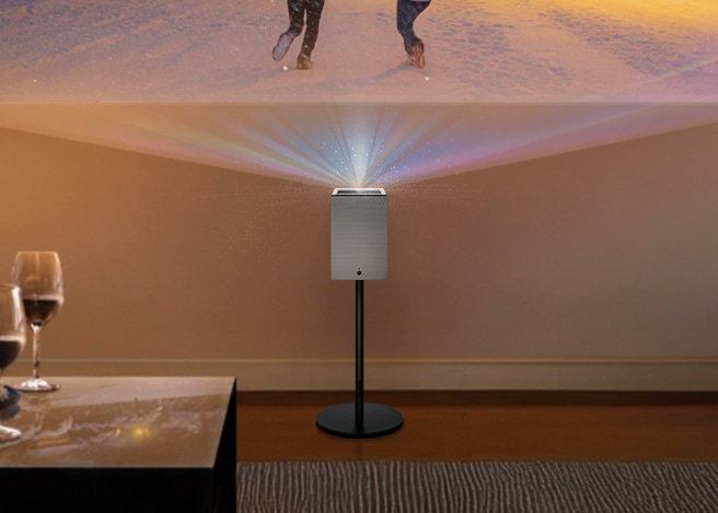 스탠드 위에 설치된 큐브 모양의 프로젝터가 정면 벽에 영상을 투사하고 있으며, 전경에는 와인잔과 테이블이 보입니다.