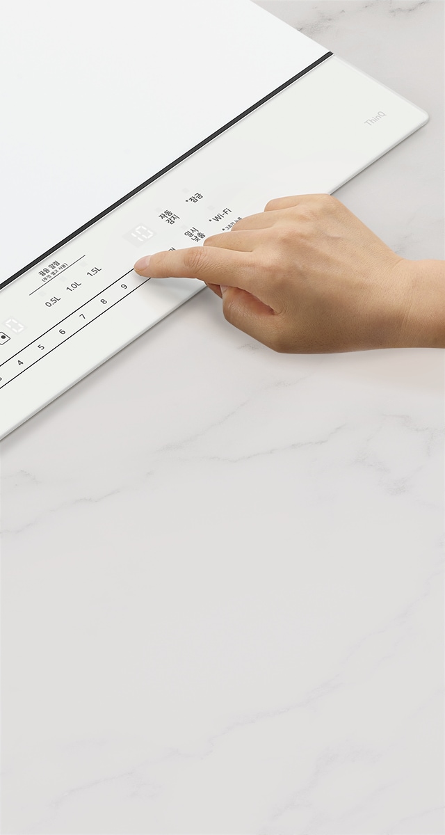 한 손이 하얀색 전자기기 패널을 터치하고 있습니다. 패널에는 최소 물량과 0.5L, 1.0L, 1.5L라는 텍스트가 있으며, 숫자 1부터 9까지의 숫자가 나열되어 있습니다. 또한 자동 급수, 인공지능, Wi-Fi 등의 텍스트가 보입니다. 패널의 한쪽에는 ThinQ라는 로고가 있습니다. 배경은 대리석 무늬의 표면입니다.