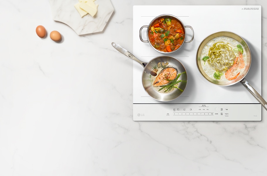 하얀색 인덕션 위에 세 개의 냄비가 놓여 있습니다. 첫 번째 냄비에는 연어와 아스파라거스가 들어 있고, 두 번째 냄비에는 다양한 채소가 들어 있는 붉은 소스 요리가 있습니다. 세 번째 냄비에는 새우와 브로콜리가 들어 있는 크림 소스 요리가 있습니다. 인덕션 주변에는 계란 두 개와 치즈 조각이 있는 대리석 도마가 있습니다. 인덕션에는 LG 로고와 다양한 조작 버튼이 보입니다.