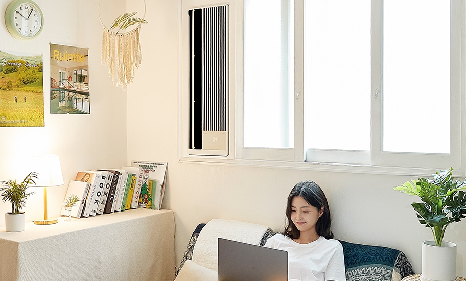 앉아서 노트북을 보고 있는 여성과 책, 조명, 화분이 놓인 탁자, 창문에 설치된 창호형 에어컨