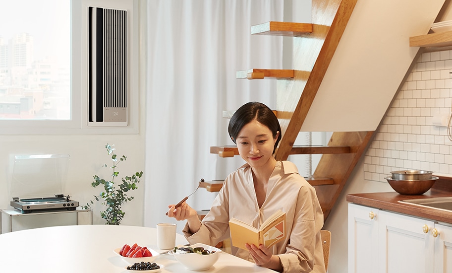 식탁에 앉아 음식을 먹으며 책을 보고 있는 여성과 뒤쪽에 설치된 창호형 에어컨